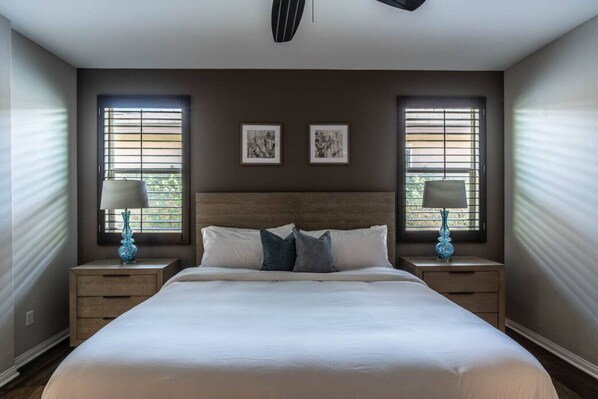 A bedroom with a large bed, two nightstands, and two windows with blinds.