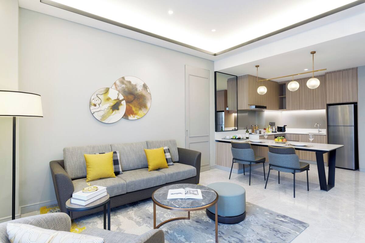Un salon moderne avec un canapé gris, des coussins jaunes, une table basse et un pouf rond. On aperçoit, au fond, une cuisine avec un coin repas.