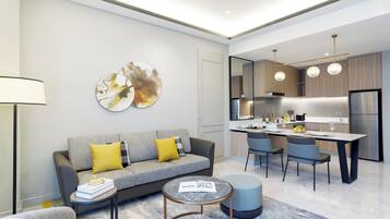 A modern living room with a grey sofa, yellow cushions, a coffee table, and a round ottoman. A kitchen with a dining area is visible in the background.