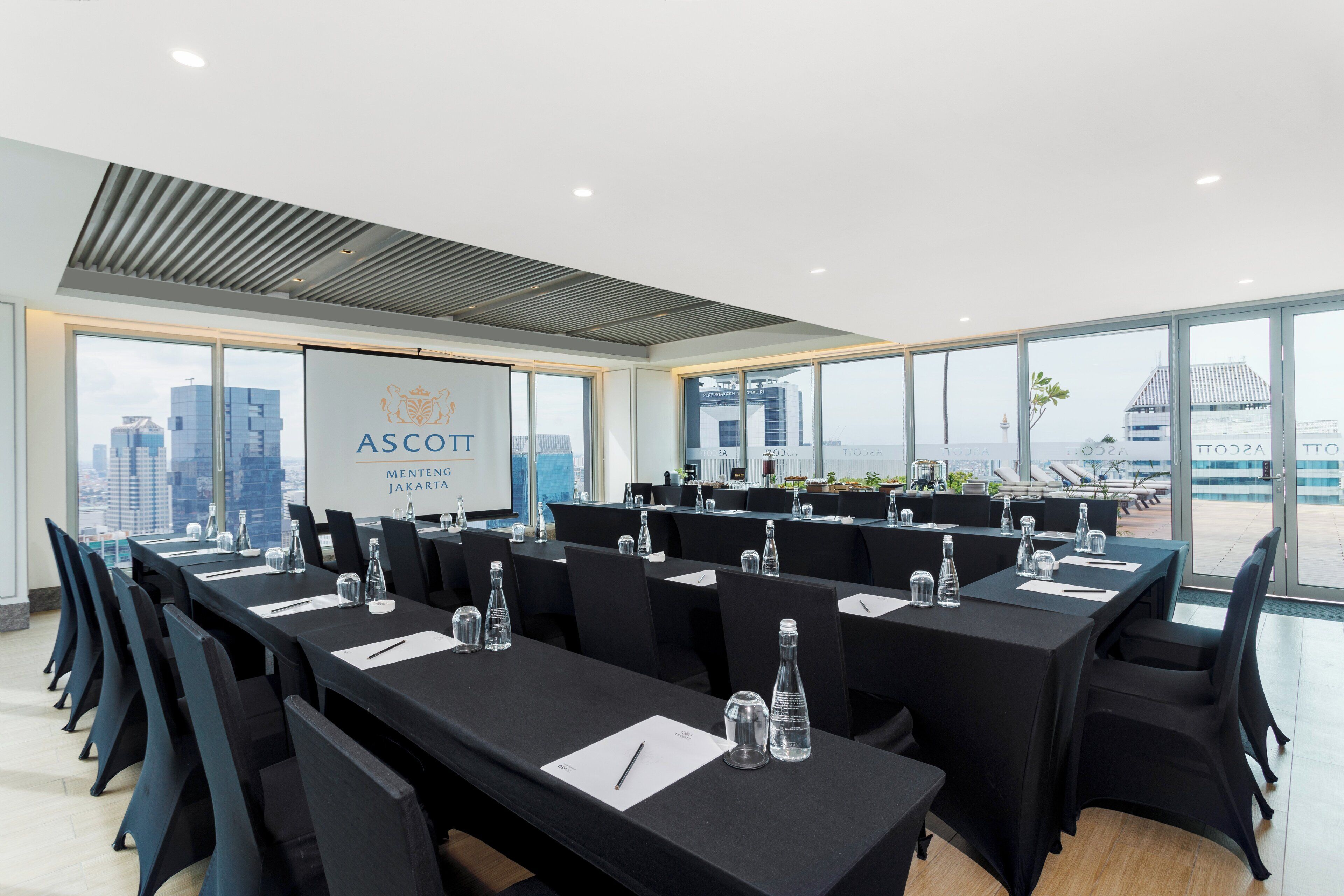Un salón de conferencias con varias mesas, sillas y una pantalla grande que muestra el logo "ASCOTT MENTENG JAKARTA".