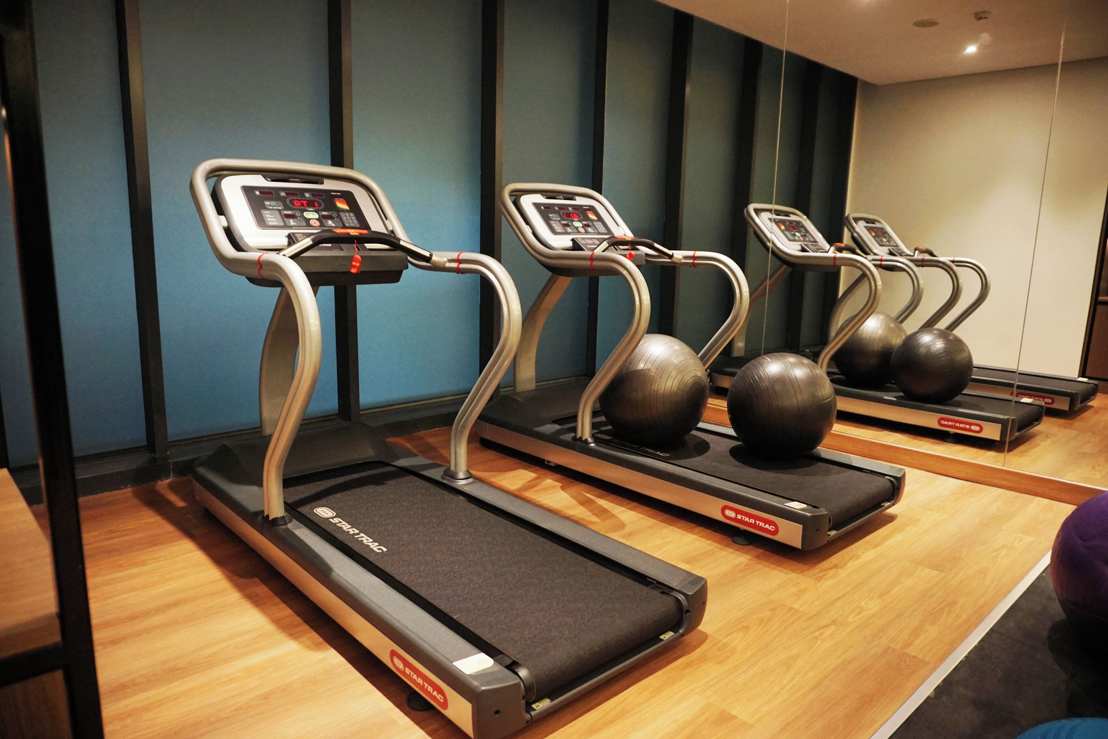 Een rij loopbanden met digitale beeldschermen, geplaatst in een sportschool met houten vloer en blauwe wanden.