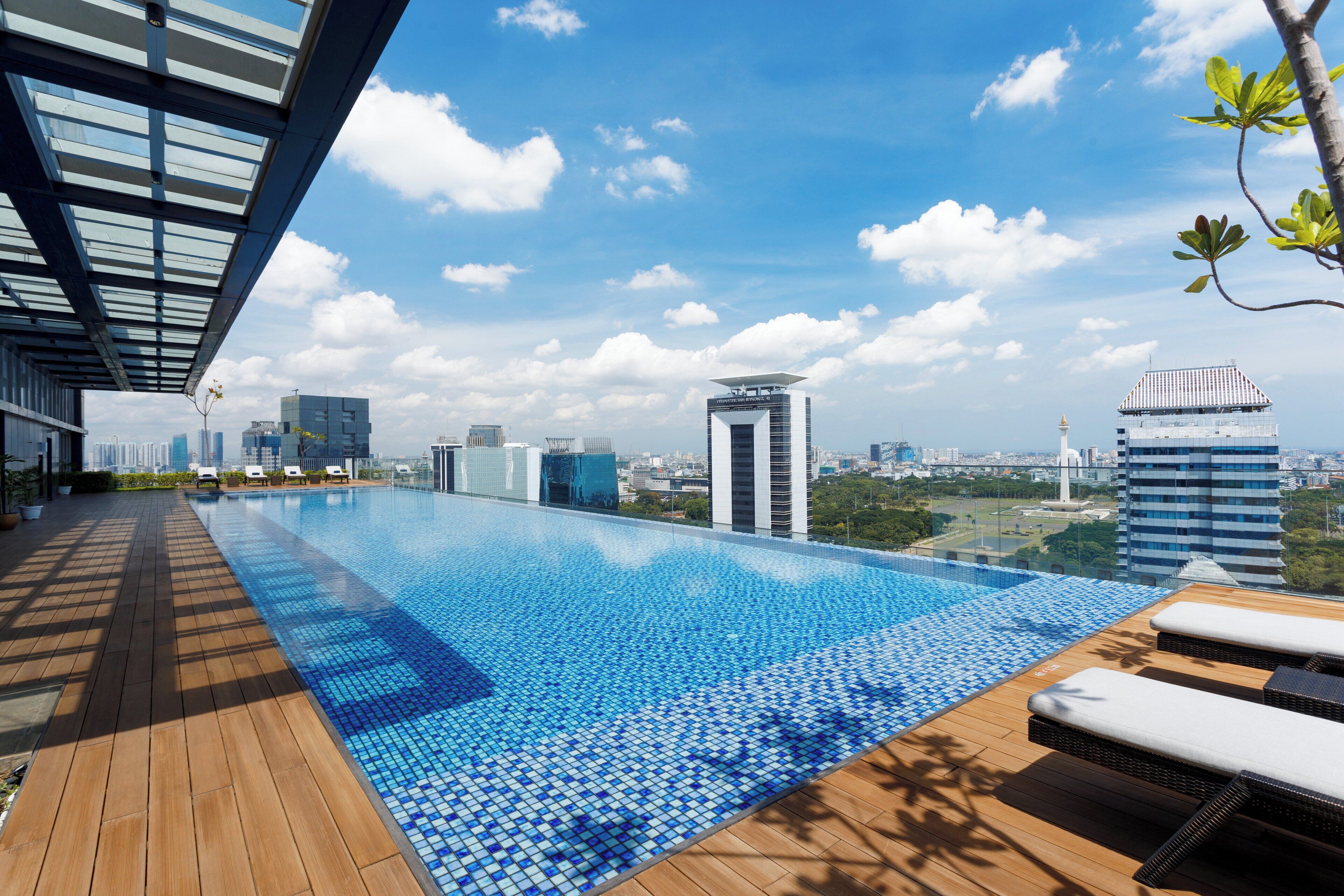 A rooftop infinity pool with wooden deck and lounge chairs, overlooking a cityscape with modern buildings and a clear sky.
