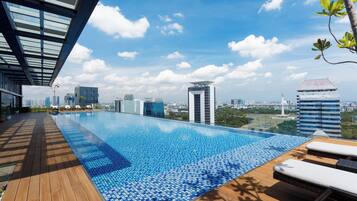 A rooftop infinity pool with wooden deck and lounge chairs, overlooking a cityscape with modern buildings and a clear sky.
