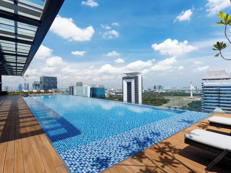 A rooftop infinity pool with wooden deck and lounge chairs, overlooking a cityscape with modern buildings and a clear sky.