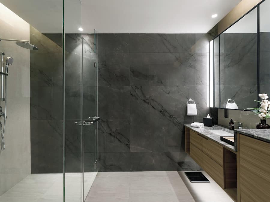 Un baño moderno con ducha envuelta en cristal, paredes de mármol, tocador de madera y un espejo grande.
