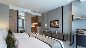 A modern hotel room with two beds, a TV, a desk, and a small kitchenette.