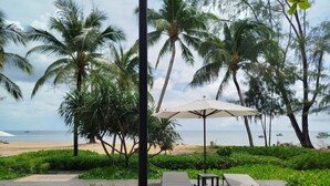 A tropical garden with a pathway leading to a beach. There are palm trees, a beach umbrella, and lounge chairs. - Kalia Phu Quoc Hotel 2 (Phu Quoc)