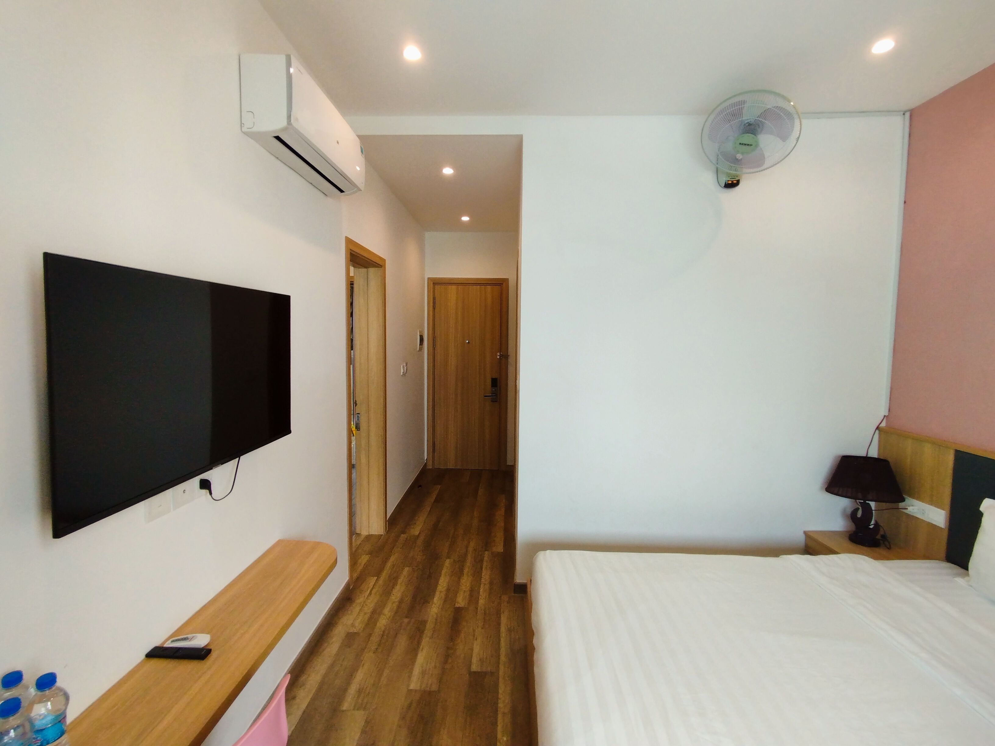 Habitación de hotel con cama, televisor de pantalla plana montado en la pared, banco de madera, mesita de noche con lámpara y ventilador de techo.
