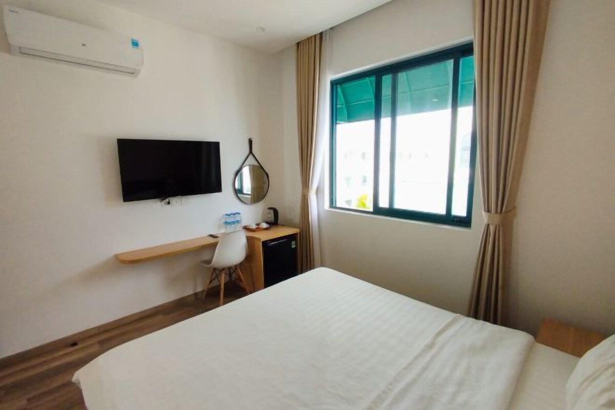 Quarto de hotel com cama, escrivaninha com cadeira, televisão e janela com cortinas.