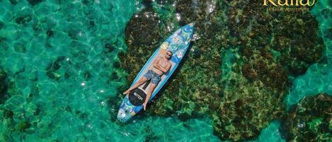 A person lying on a surfboard in clear turquoise water, surrounded by coral reefs.