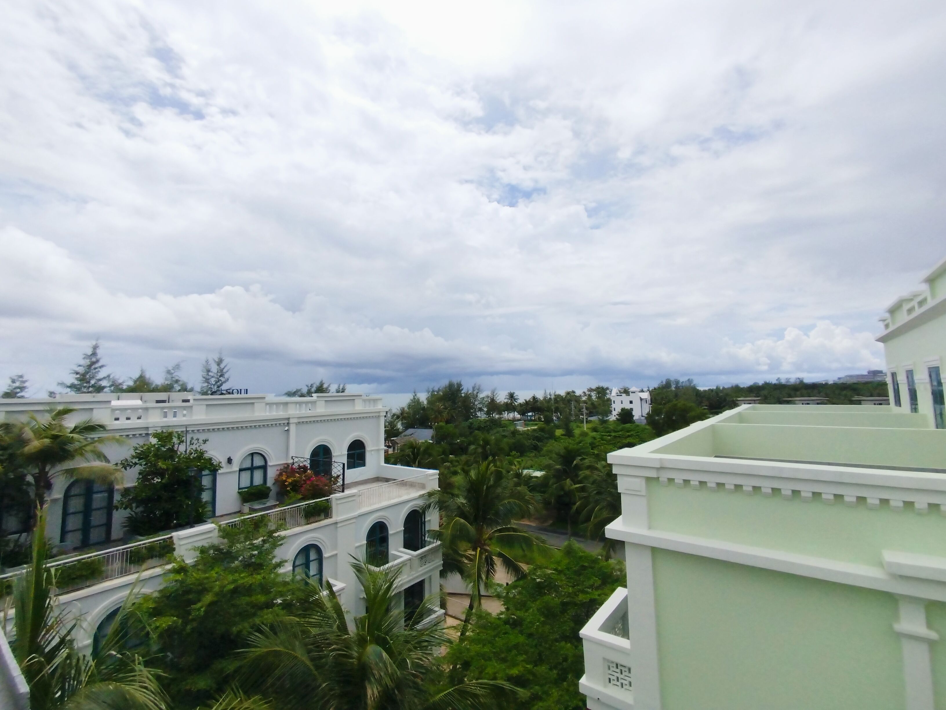 Une vue d’un quartier résidentiel avec des bâtiments blancs, une végétation luxuriante et un ciel nuageux.