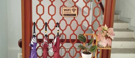 Una puerta de madera con un diseño geométrico, un cartel con la contraseña de WiFi, y un perchero con sombrillas y plantas en macetas.
