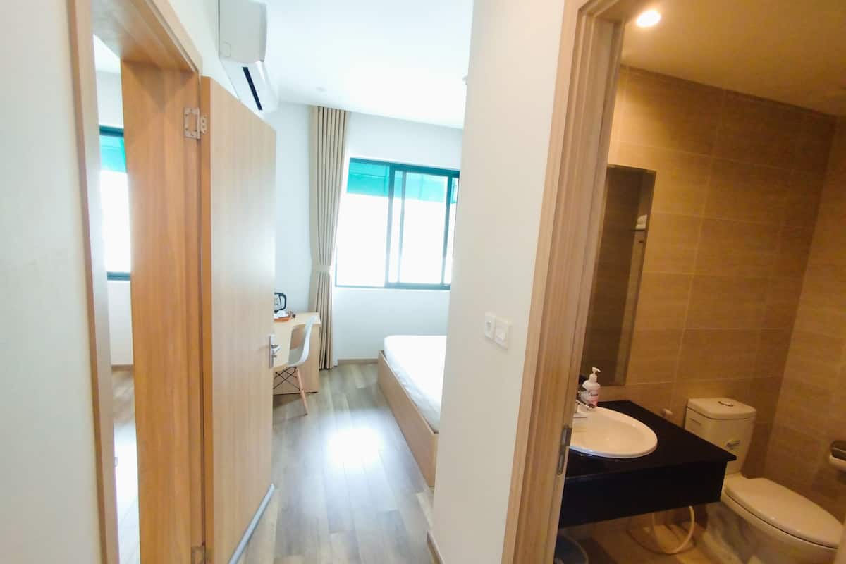 Camera d'albergo compatta con un letto, una scrivania con sedia, una finestra con tende, un bagno con lavandino e servizi igienici, e un cestino.