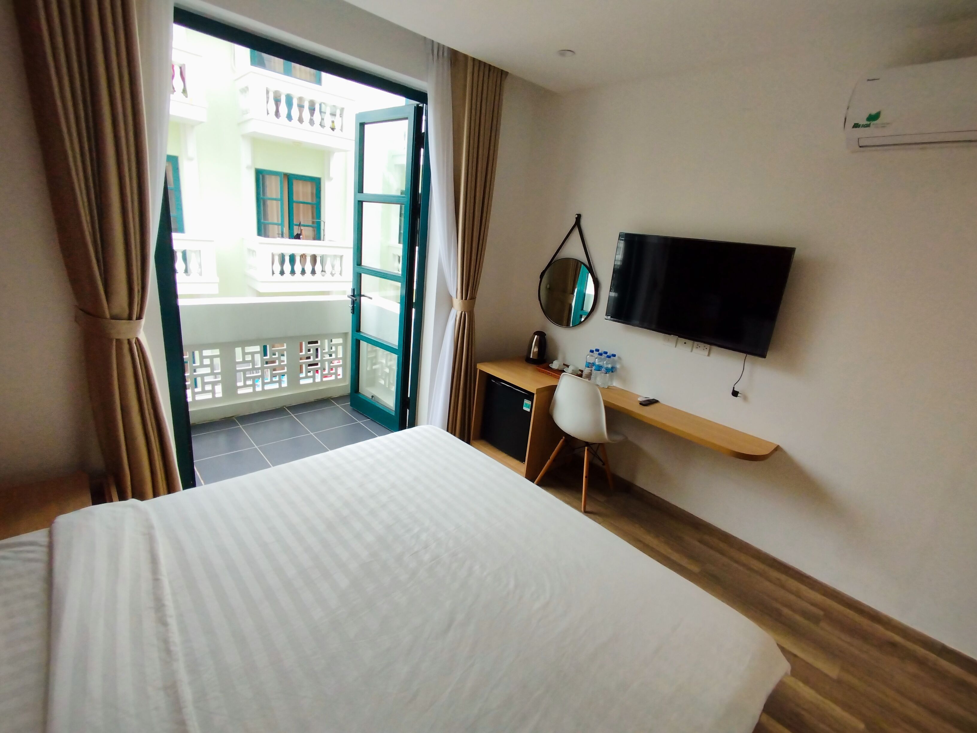 A hotel room with a bed, a desk with a chair, a TV, and a balcony with a view of buildings.