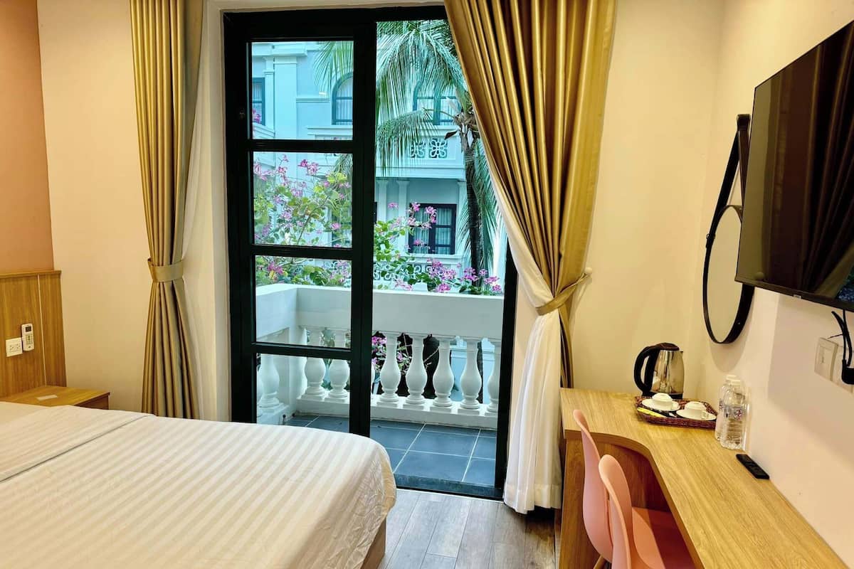 Una camera d'albergo con un letto, una scrivania con sedia, una televisione e un balcone con vista su un edificio e spazi verdi.