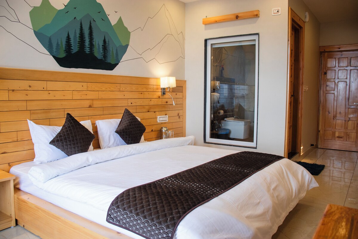 Habitación de hotel con cabecera de madera, cama grande con ropa blanca y una colcha marrón acolchada, lámpara de pared y puerta que da a un baño.