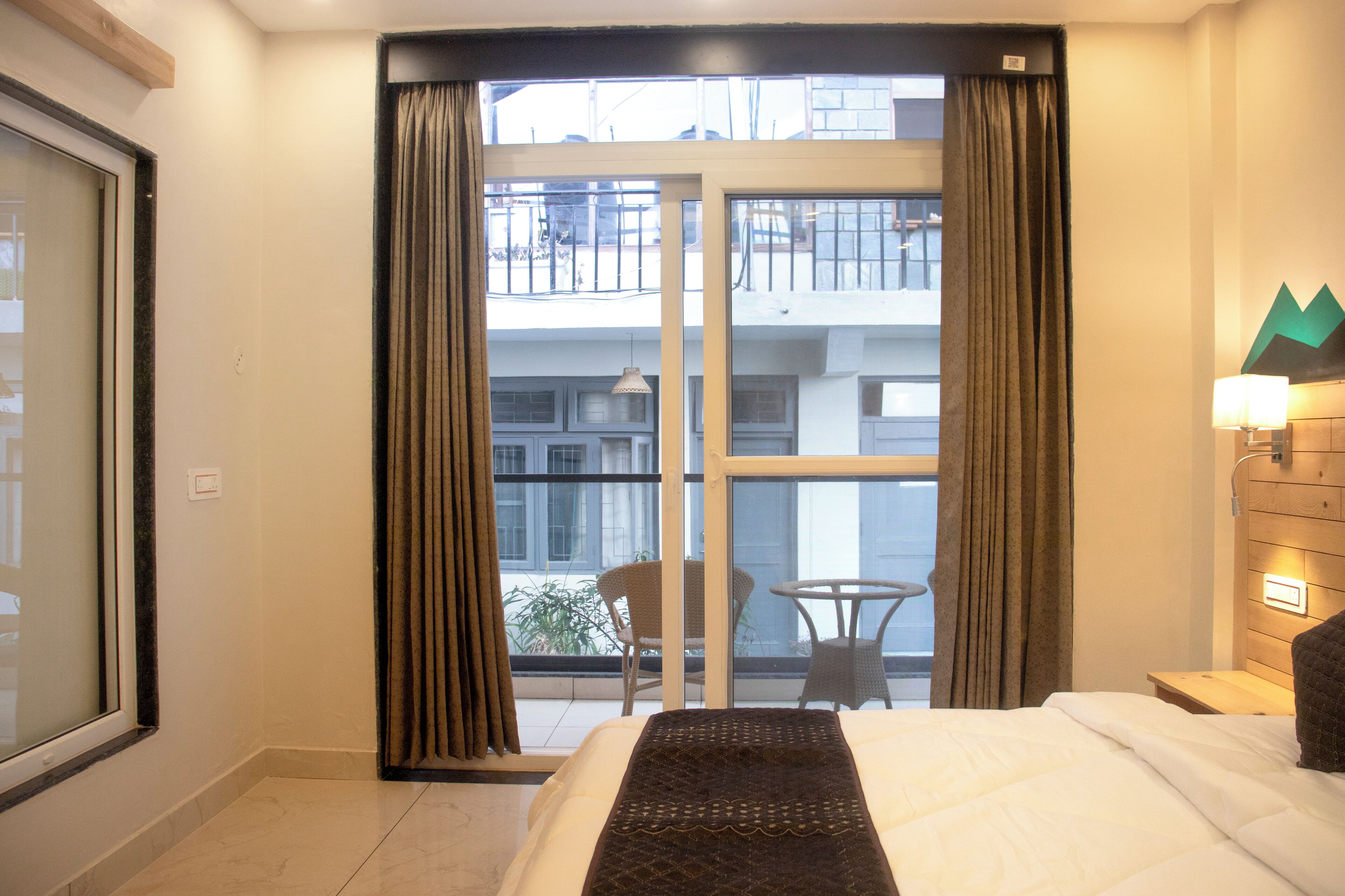 A hotel room with a bed, a window with curtains, a balcony with a chair and table, and a wall-mounted lamp.