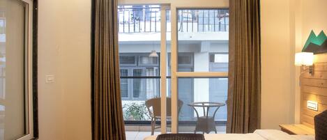 A hotel room with a bed, a window with curtains, a balcony with a chair and table, and a wall-mounted lamp.