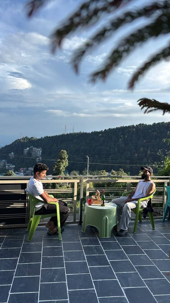 Deux personnes sont assises sur des chaises vertes, sur une terrasse d’où l’on peut admirer un paysage vallonné.