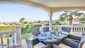 A covered outdoor dining area with a glass table and wicker chairs, overlooking a landscaped garden and distant buildings. - Spectacular villa with private pool and sea views in Denia (Denia)