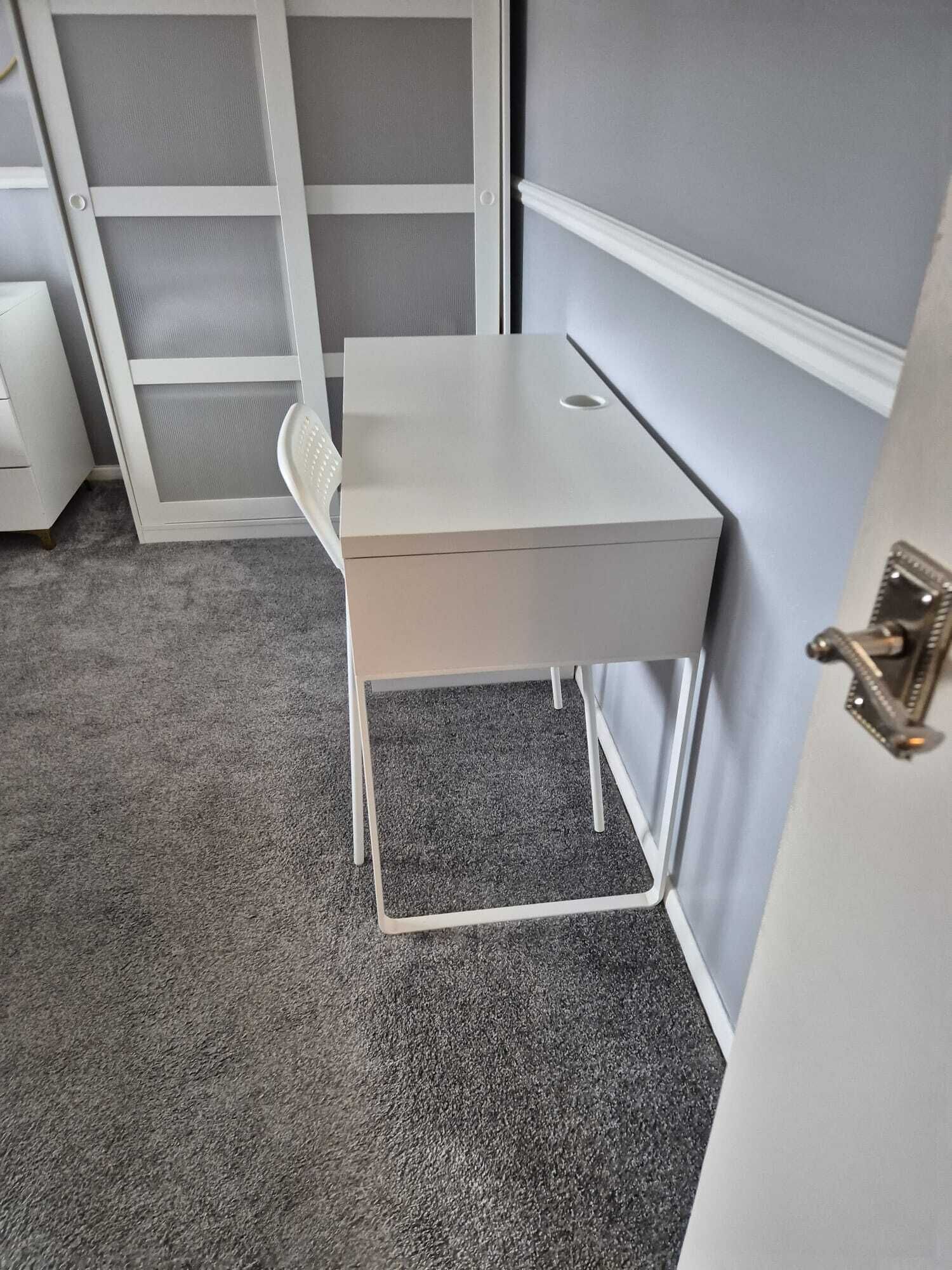 A small, white desk with a drawer, positioned against a wall with a grey door and a white door frame.