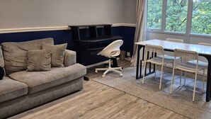 Un salon comprenant un canapé, une table à manger avec des chaises, un meuble de rangement noir pour appareils électroniques et une grande fenêtre donnant sur la forêt.