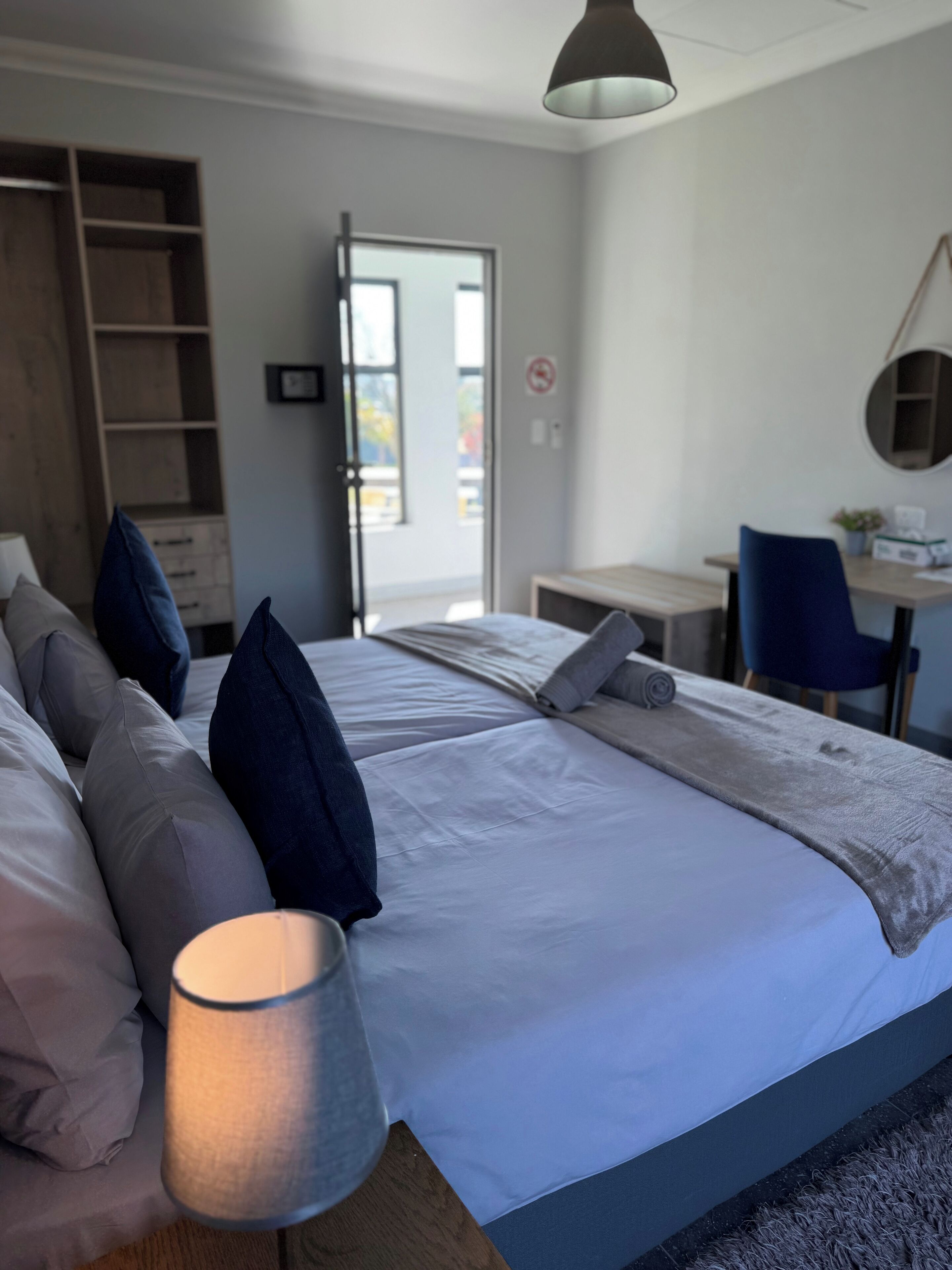 Un lit bien fait, avec des oreillers, une lampe de chevet, un bureau avec une chaise, et une vue sur l’extérieur par une porte.