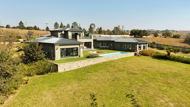 Una casa moderna de dos pisos con piscina, muros de contención de piedra y un molino de viento al fondo.
