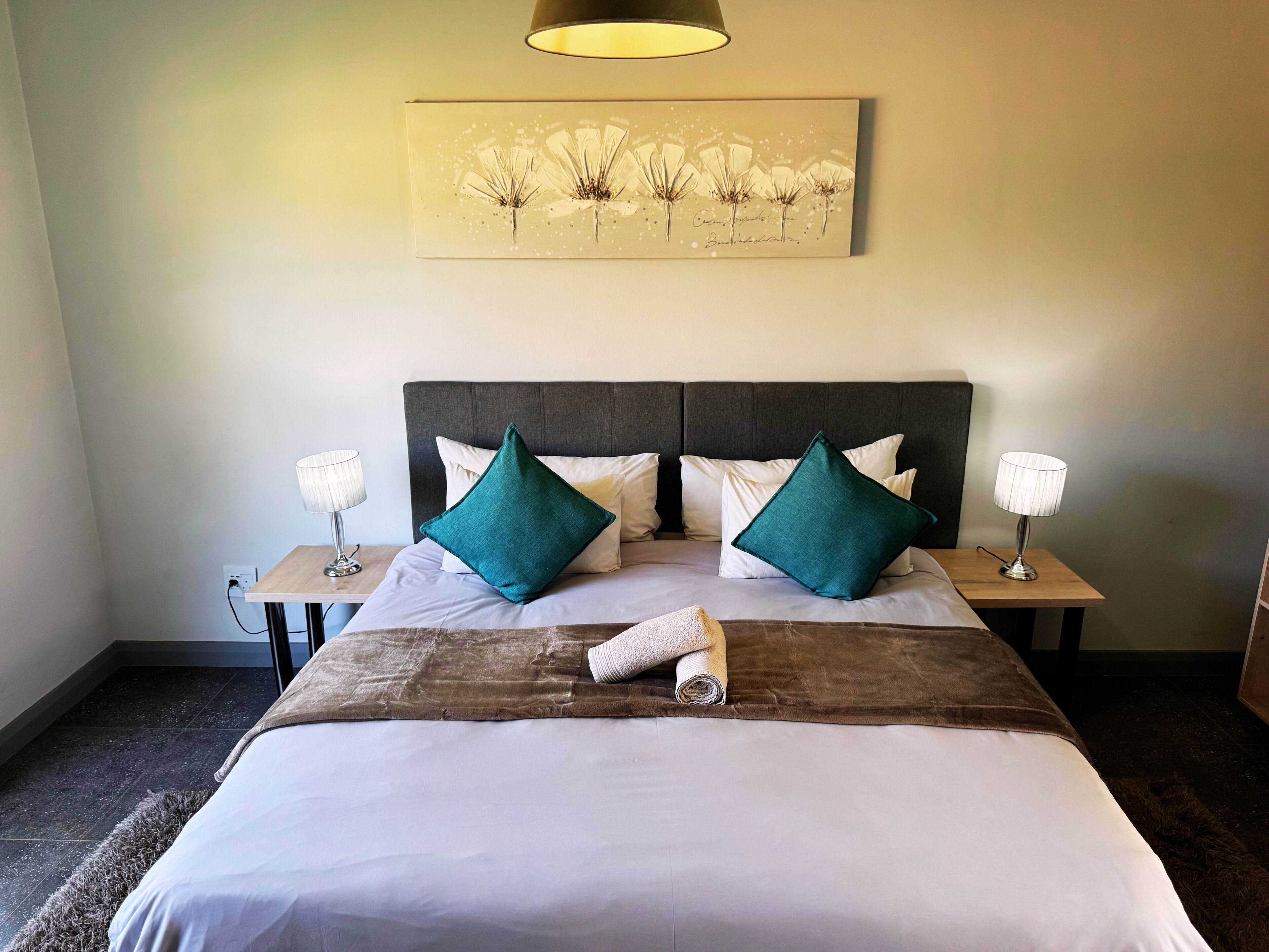 Un lit bien fait, avec une tête de lit marron, deux oreillers blancs, deux oreillers turquoise et une serviette enroulée. Il y a une table de chevet avec une lampe de chaque côté du lit. Une œuvre d’art encadrée est accrochée au-dessus du lit.