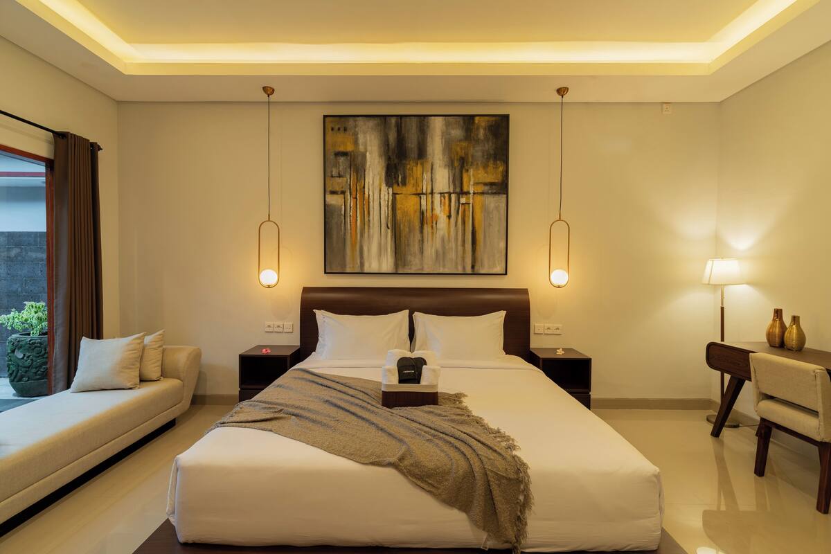 A modern hotel room with a large bed, a sofa, a desk, and a painting on the wall.