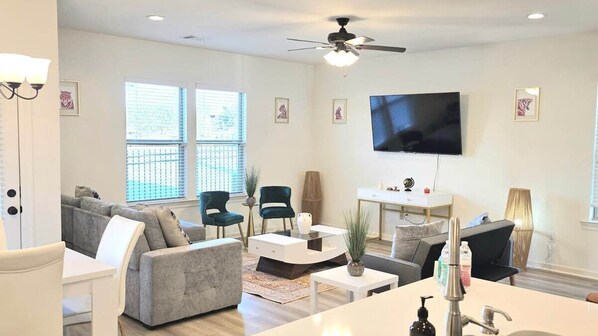 A modern living room with a flat-screen TV, a ceiling fan, and a large window.