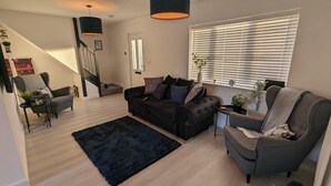 A modern living room with a black sofa, grey armchair, and a black rug. There is a staircase with a black railing, a black pendant light, and a window with blinds. - Modern Gilliver house Rural retreat sleeps 7 (Newbold Verdon)