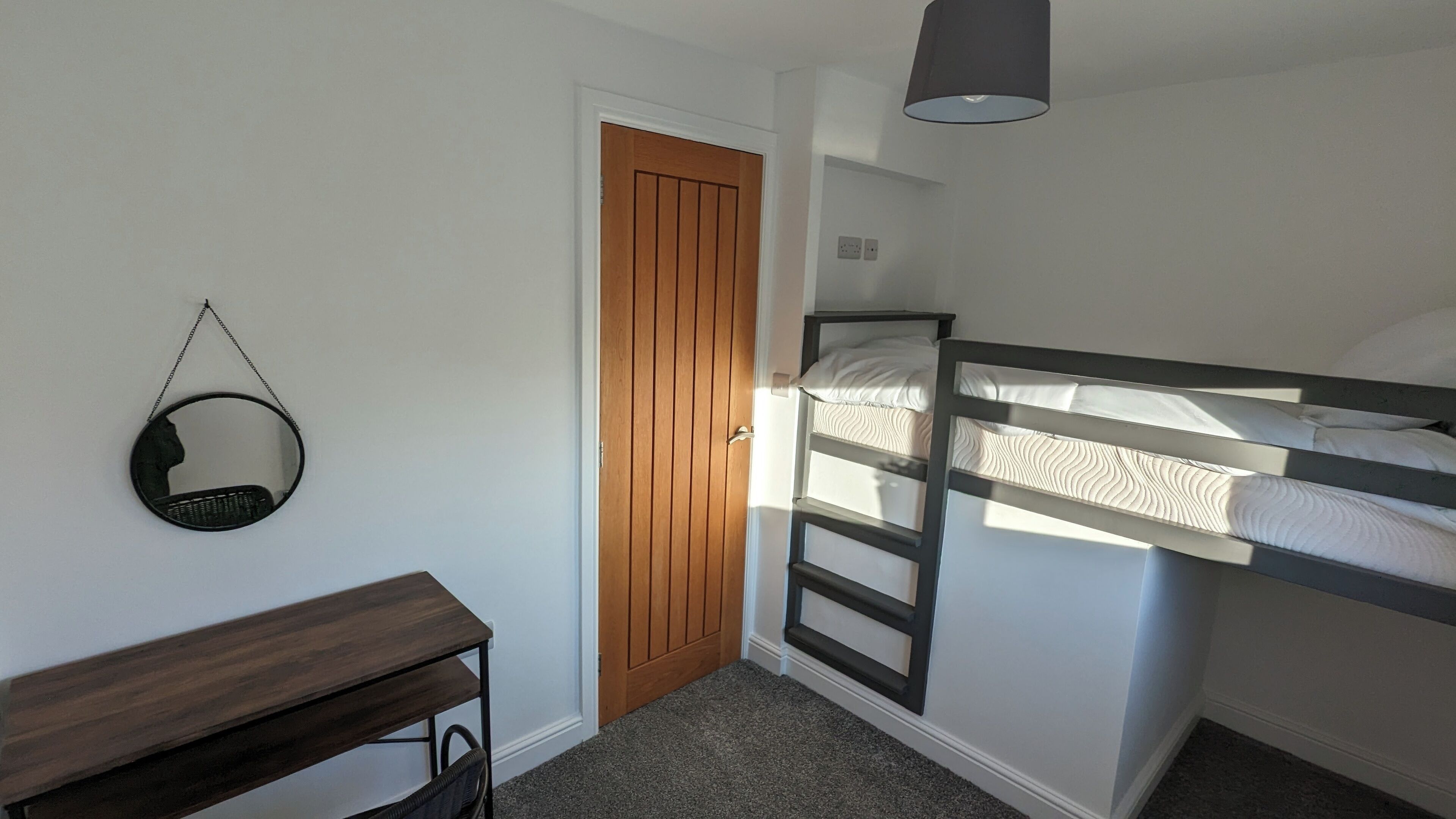 A bedroom with a bunk bed, a wooden door, a round mirror, a desk, and a lamp.