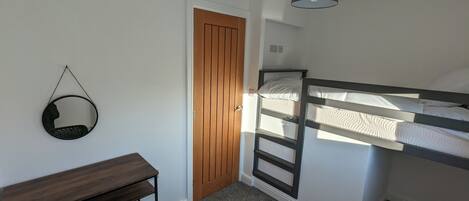 A bedroom with a bunk bed, a wooden door, a round mirror, a desk, and a lamp.