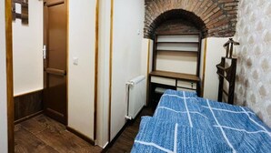 A bedroom with a wooden ceiling, a brick fireplace, a bed with a blue and white patterned bedspread, a wooden wardrobe, and a radiator.