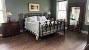Un dormitorio con una cama grande, piso de madera, dos mesitas de noche, un aparador, un ventilador de techo y un espejo amplio.