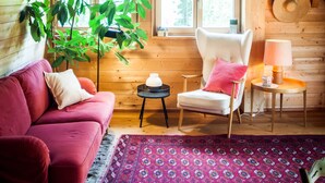 Una habitación acogedora con un sofá rojo, una silla blanca, una mesita de noche con una lámpara, y un ventanal con vistas a árboles.