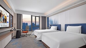 Une chambre d’hôtel avec deux lits, un bureau et une chaise. Elle dispose d’une grande fenêtre donnant sur la ville.