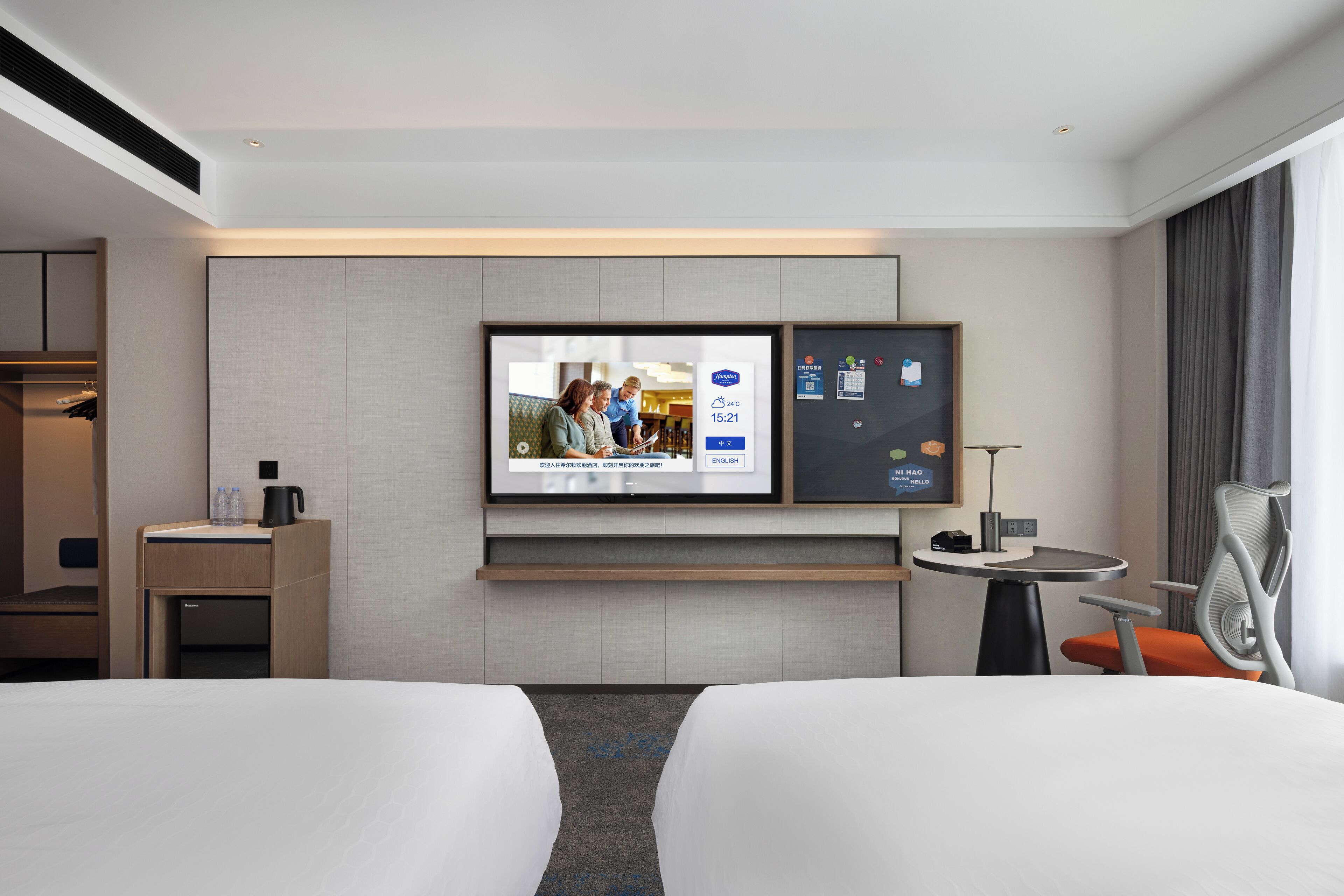 A hotel room with a bed, a TV mounted on the wall, a desk with a chair, and a small table with a lamp.