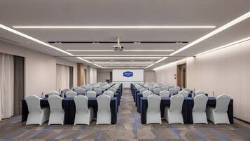 A conference room with rows of tables and chairs, a projector screen, and a carpeted floor.