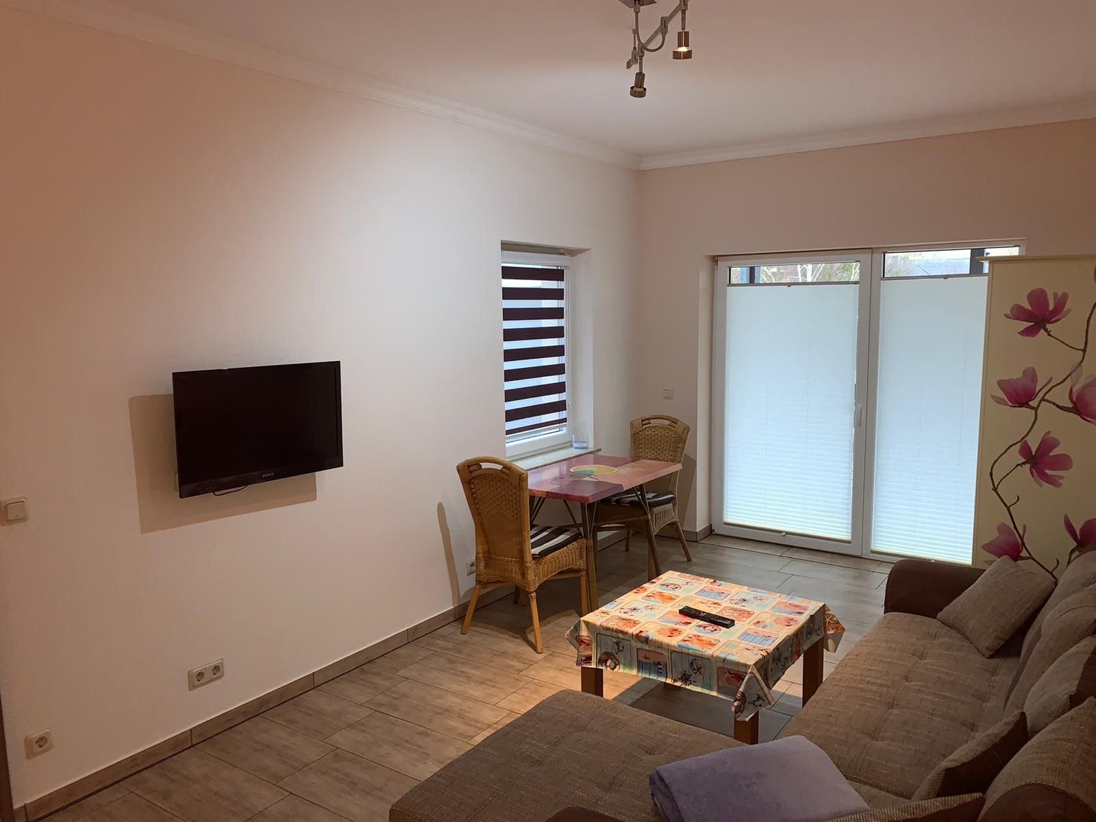 Un salon avec un téléviseur à écran plat fixé au mur, une table à manger avec des chaises, un canapé et une fenêtre avec des stores.