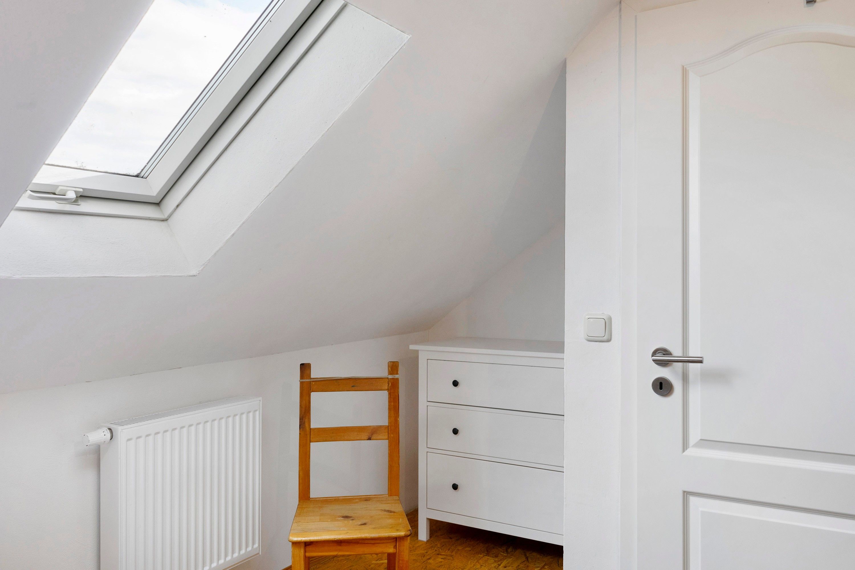 A room with a sloped ceiling, a radiator, a wooden chair, a white dresser, and a door with a lock.
