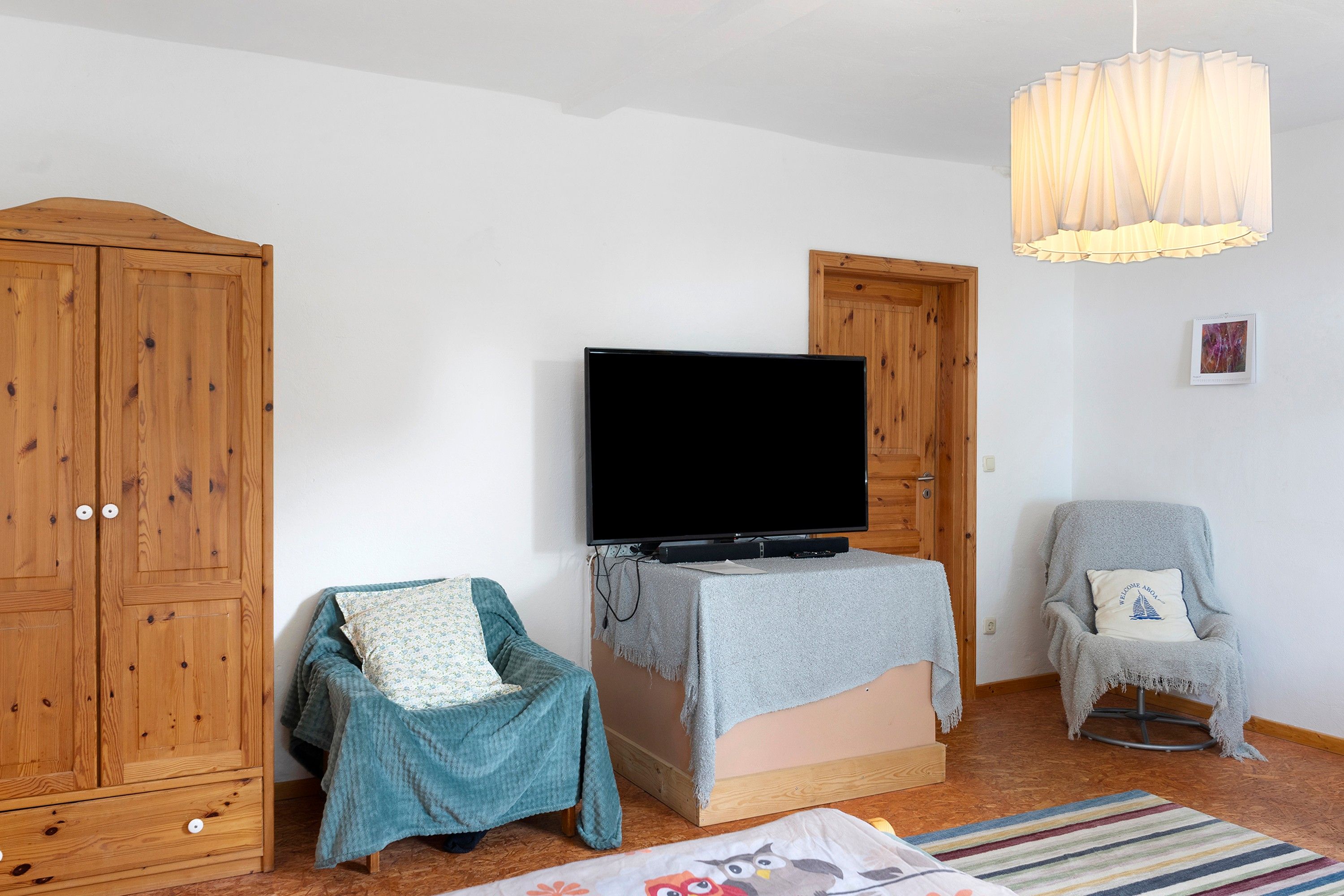 Ein Zimmer mit Fernseher auf einem Schrank, einem Holzschrank, einem kleinen Sessel mit Kissen und einer Pendelleuchte.