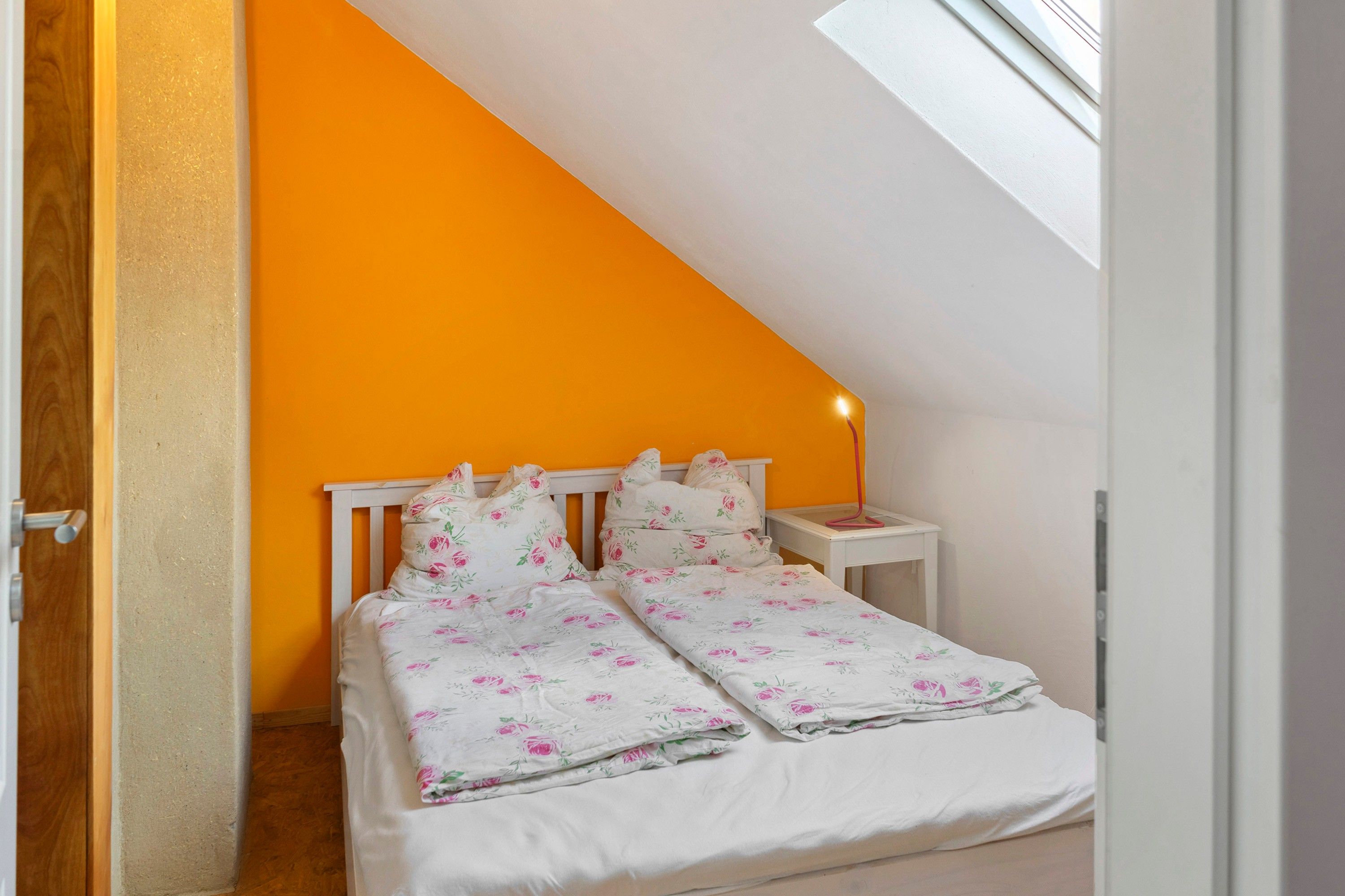 Un dormitorio con una pared naranja, una cama blanca con ropa de cama floral, una mesita de noche con una lámpara y un tragaluz.