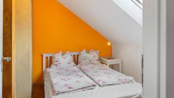 Un dormitorio con una pared naranja, una cama blanca con ropa de cama floral, una mesita de noche con una lámpara y un tragaluz.