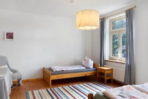 Et børneværelse med en enkel seng, et lille skrivebord, en stol, et vindue med gardiner og en hængende lampe.