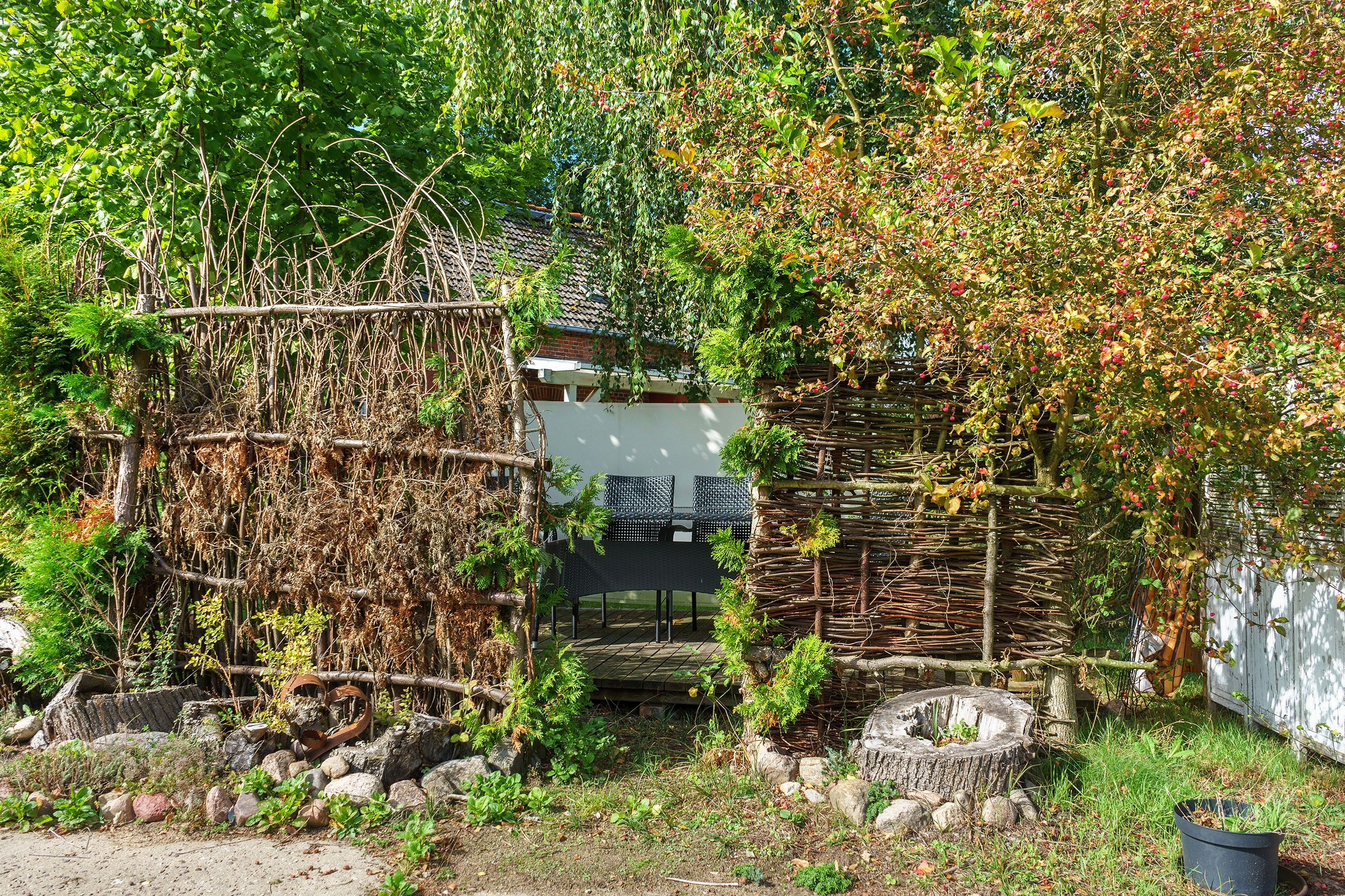 Ein rustikaler Außenbereich mit einer Holzkonstruktion, einer weißen Wand und einem Garten mit verschiedenen Pflanzen und Steinen.