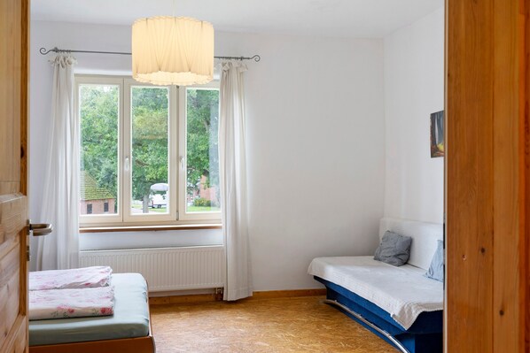 A small room with a window, a radiator, a bed, and a wooden door.