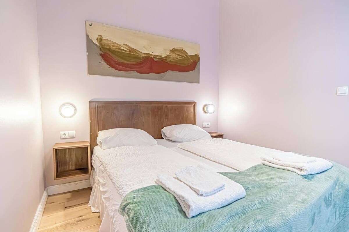 A double bed with white linens, a green throw blanket, and folded towels on the bed. There is a wooden headboard, two bedside tables with lamps, and a wall-mounted artwork above the bed.