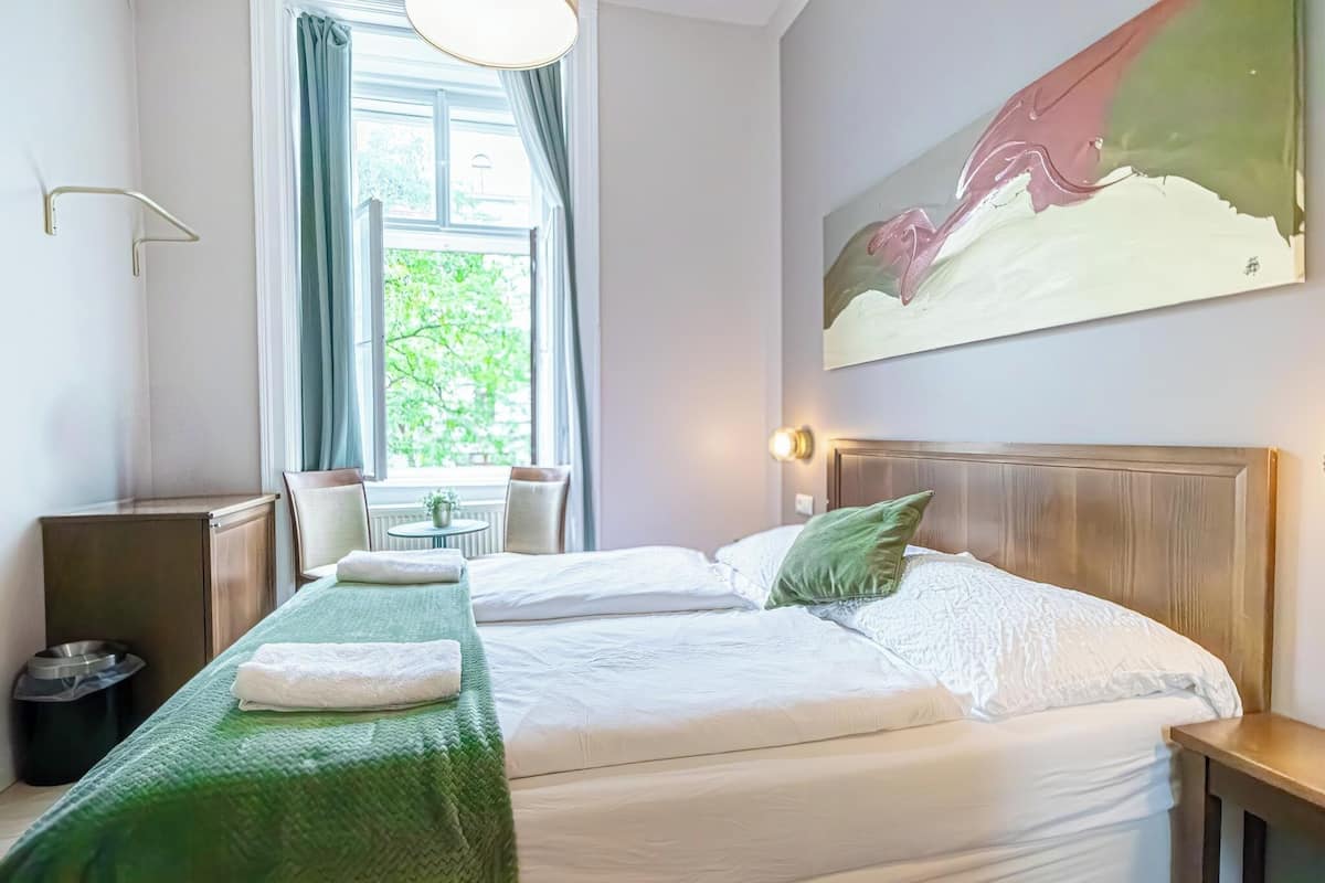 A hotel room with two beds, a window with curtains, a painting on the wall, and a small table with chairs.